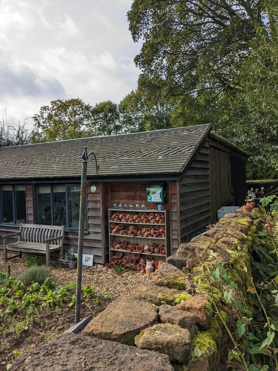 Single-storey timber-clad garden building with a tiled roof, a bench and shelves of terracotta pots, behind a dry-stone wall