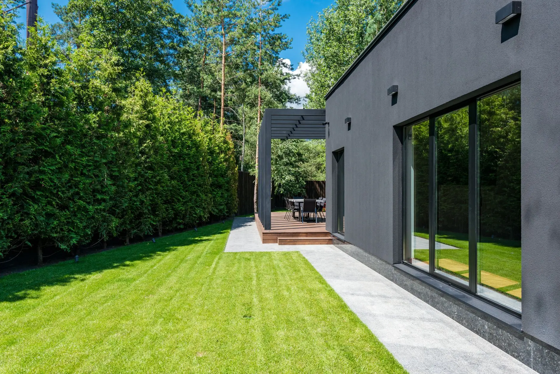 Single-storey building with full-height sliding glass doors opening onto a striped lawn, with a tall hedge alongside