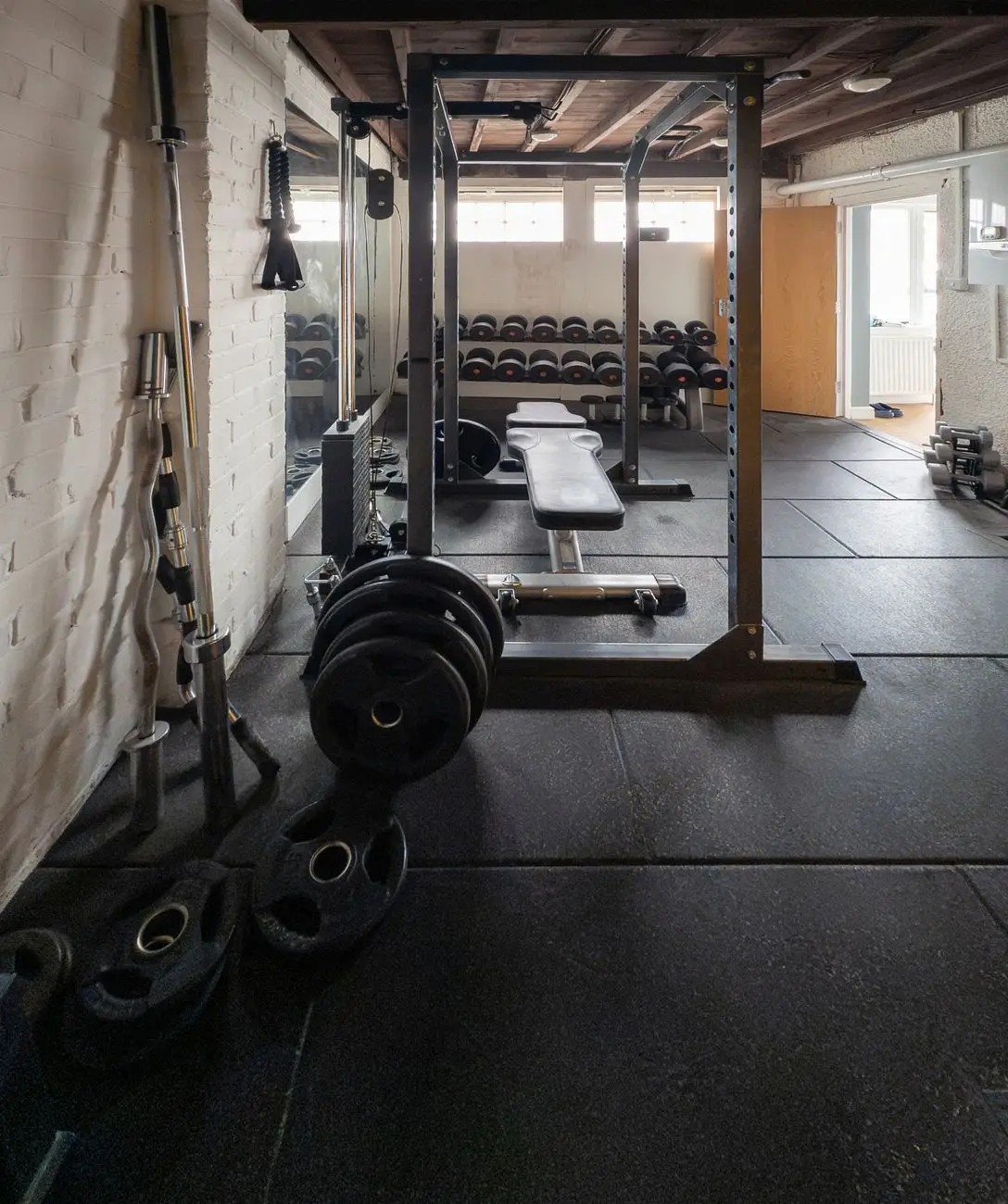 Home gym with a squat rack, weight plates, a bench and rubber floor tiles against a painted brick wall