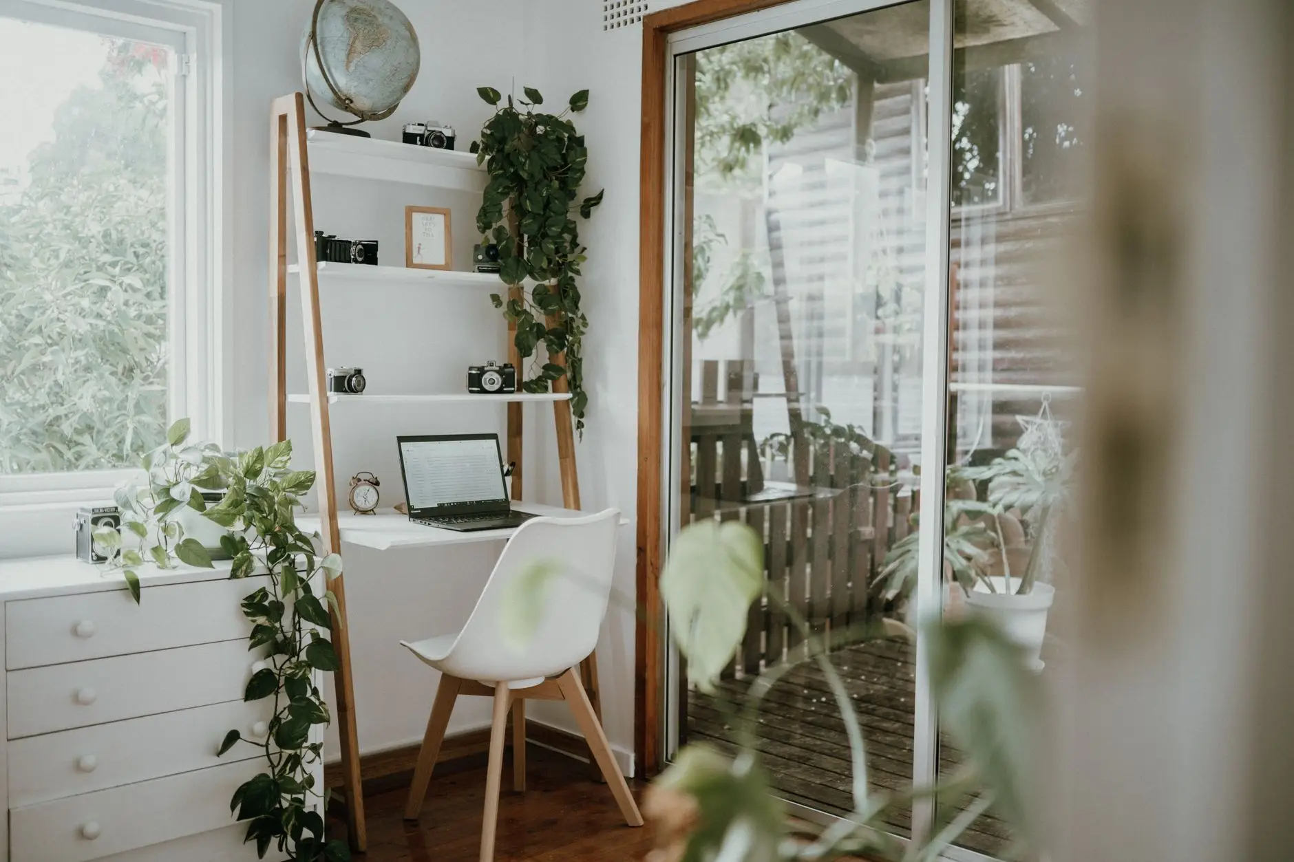 Bright home office desk with a laptop, shelving and plants beside glazed doors