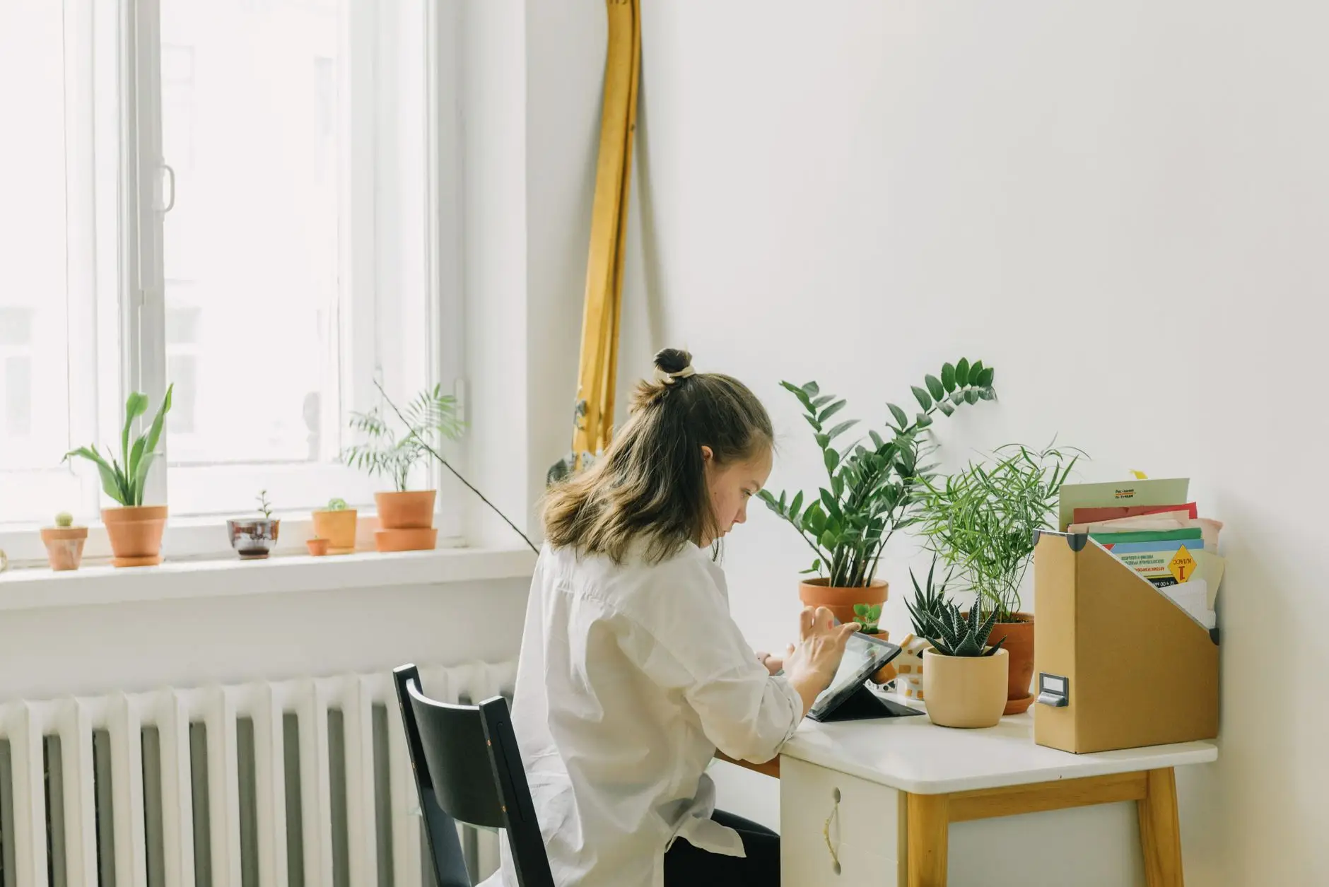 Woman working at a small desk by a window, with potted plants and a white radiator along the wall